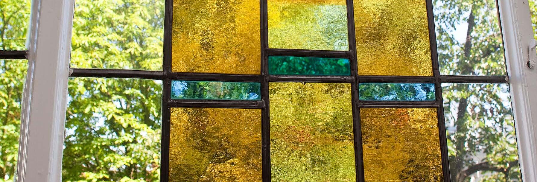 Bild zeigt eine Nahaufnahme eines mit buntem Glas verziertes Bleiglasfenster aus dem späten 19. Jahrhundert. In der Mitte zeigt eine Malerei eine zieselierte Empore, in dessen Blumenkübel unterschiedliche Blüten sichtbar sind.