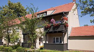 Außenaufnahme des Wohnhauses Ziegenorter Pfad. Zu sehen ist der EIngangsbereich des Hauses mit begrüntem Vorgarten und hervorspringenden, nachträglich angestellten Balkonen.