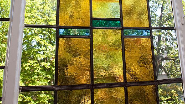 Bild zeigt eine Nahaufnahme eines mit buntem Glas verziertes Bleiglasfenster aus dem späten 19. Jahrhundert. In der Mitte zeigt eine Malerei eine zieselierte Empore, in dessen Blumenkübel unterschiedliche Blüten sichtbar sind.