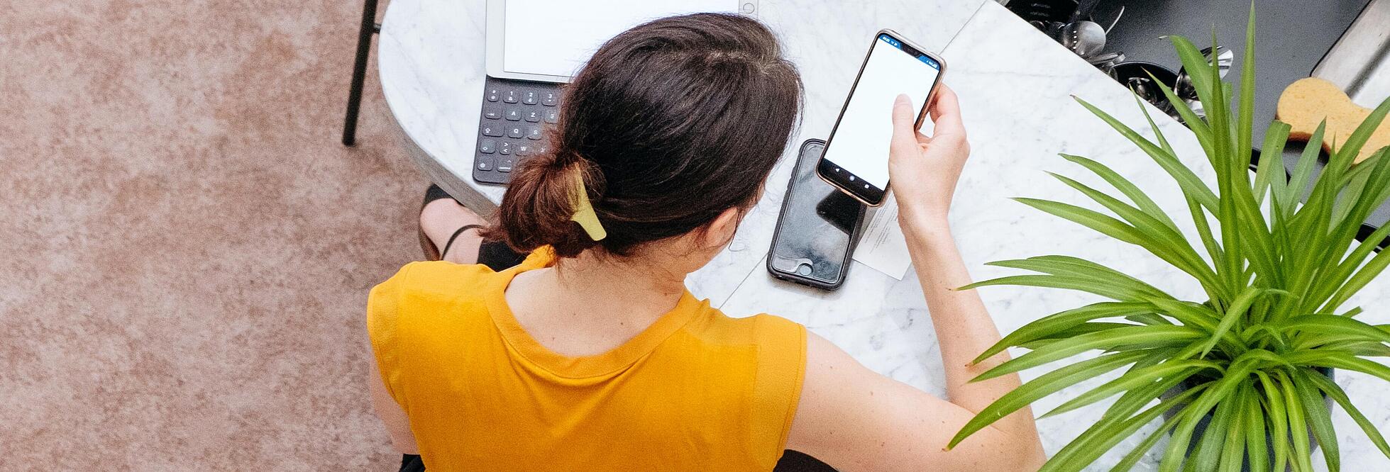 Foto von oben: Eine Frau sizt mit dem Rücken zur Kamera an einem L-förmigen, schmalen Tisch und bedient ein Smartphone. Unter diesem ist ein weiteres sichtbar, links verdeckt von ihrem Haar sieht man die Tastatur eines Laptops.